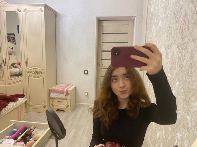 LiliaBilka mit welligem, braunem Haar, trägt ein schwarzes Langarmshirt und macht ein Selfie vor einem Spiegel, auf dem ihr Gesicht und ihre kleinen Brüste zu sehen sind. Sie hält ein rotes Telefon..
