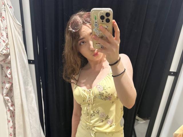 LiliaBilka, con cabello castaño ondulado, vestida con un vestido amarillo con estampado floral y botones delanteros, se toma un selfie frente a un espejo en una tienda, mostrando sus senos y labios fruncidos..