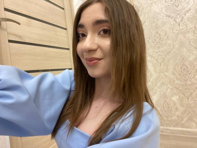 LiliaBilka, con lunghi capelli castani, indossa una camicia azzurra, sorride dolcemente alla fotocamera, mostrando il naso e le labbra, con un trucco leggero e una pelle naturale..