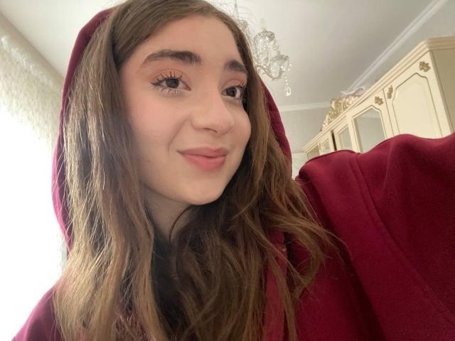 LiliaBilka, con cabello castaño largo, vistiendo una sudadera roja, sonriendo suavemente, con ojos marrones, maquillaje ligero, labios rosados y una apariencia linda y relajada..