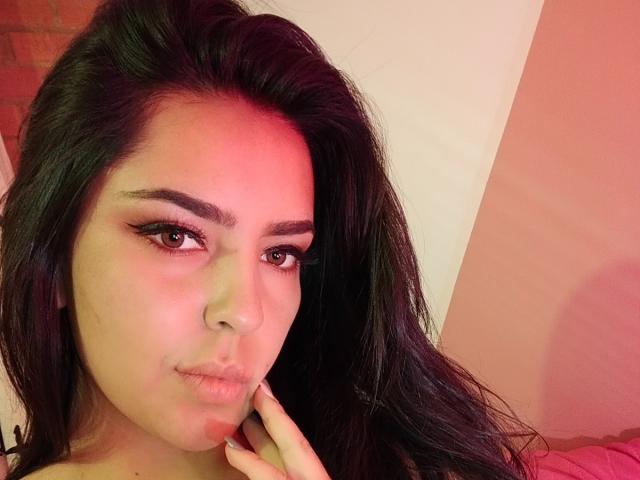 LaurynBm tiene el cabello largo y negro, ojos marrones con delineador alado, y una ligera expresión de labios fruncidos. Su mano toca su mejilla, con iluminación roja y rosa en su rostro..