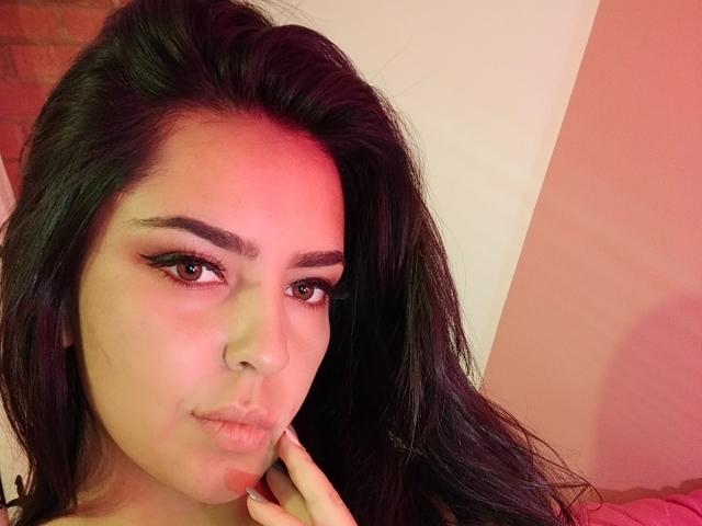 LaurynBm tiene el cabello oscuro y ondulado, labios carnosos y una ligera sonrisa, con un dedo cerca de su barbilla. Tiene cejas pobladas, pestañas largas y una luz roja que ilumina su rostro..