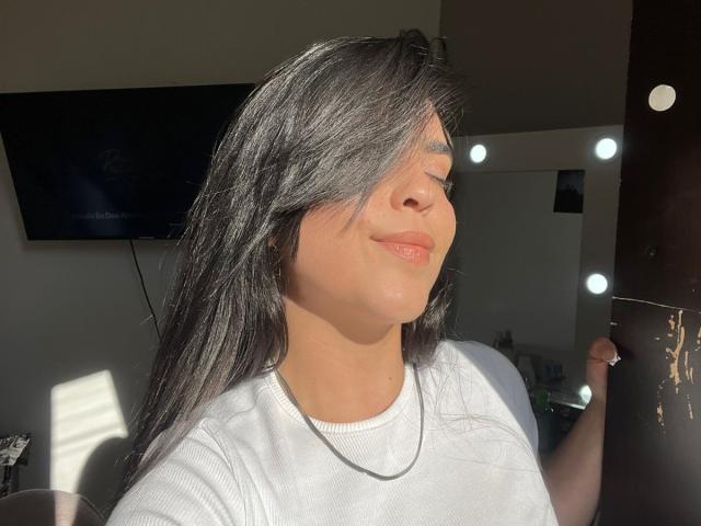 LaurynBm a de longs cheveux noirs, les yeux fermés et un léger sourire, elle porte une chemise blanche et se tient au soleil, touchant un mur sombre avec des lumières rondes..
