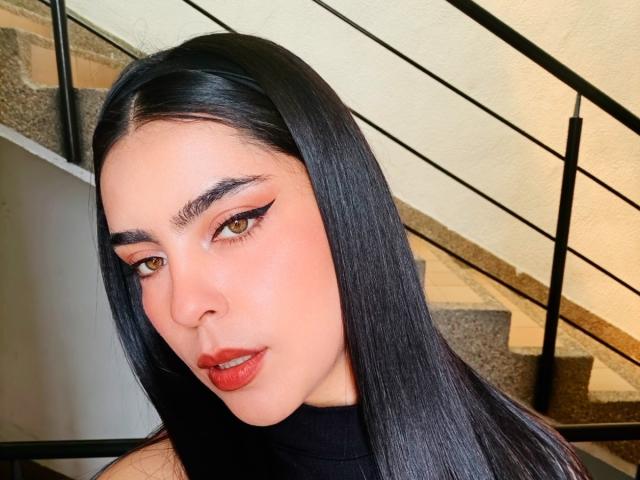 LaurynBm has long black hair, brown eyes, and red lipstick. She's wearing black eyeliner and a black turtleneck. Her ass is not visible. Stairs in background.