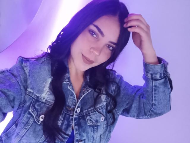 LaurynBm avec de longs cheveux noirs tressés, portant une veste en jean, une main dans les cheveux, éclairage violet-bleu, souriant doucement, montrant un visage mignon, peau claire, petit nez..