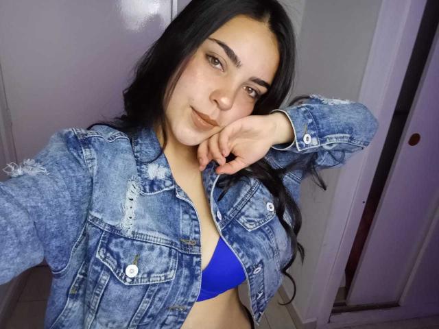 LaurynBm with long black hair, wearing a blue denim jacket, and a blue bra, resting her chin on her hand, smiling softly, showing her tits.