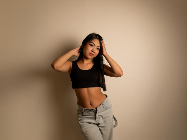 AlinnaSans mit langen, schwarzen Haaren, kleinen Brüsten und einem flachen Hintern, trägt ein schwarzes Crop-Top und hellgraue Hosen und steht vor einer beige farbenen Wand, die Hände im Haar..