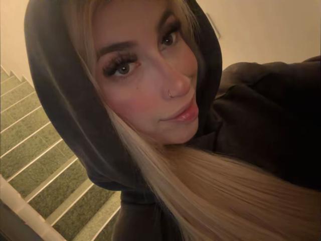SammyViolet, com longos cabelos loiros, vestindo um moletom escuro, com um piercing no nariz e delineador marcante, olhando para a câmera, sorrindo levemente, com o traseiro quase imperceptível ao fundo, nas escadas..