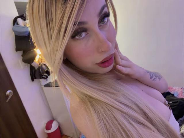 SammyViolet, avec ses longs cheveux blonds, son maquillage complet et son rouge à lèvres rouge, regarde l'appareil photo, touche sa joue, montre ses seins volumineux, a un tatouage sur son bras droit, dans une chambre..