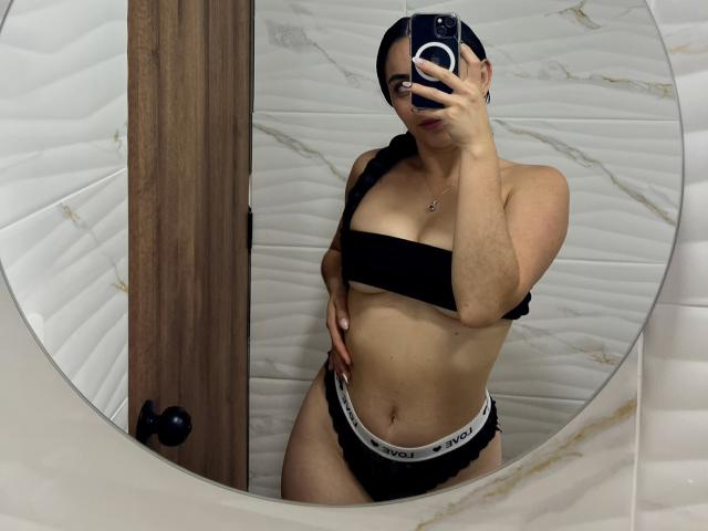 AllisonTasty, con un top de tirantes negro y ropa interior de Victoria's Secret, de pie frente a un espejo, sosteniendo un teléfono, mostrando sus pechos y glúteos, con una trenza y gafas..