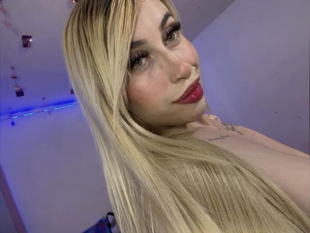 SammyViolet tem cabelos loiros longos, batom vermelho e um piercing no nariz. Ela está olhando para a câmera com um leve sorriso, mostrando suas tatuagens e seios grandes..