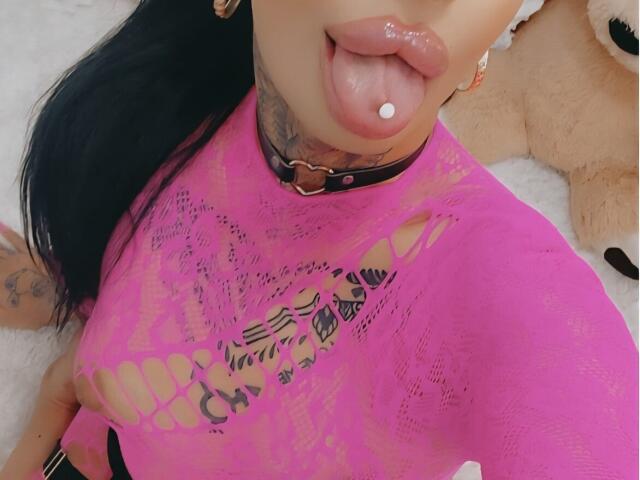 DenyChantal with black hair, tongue out, tongue piercing, pink lace top, tattoos, black choker, visible cleavage, soft pink lips, on white carpet, stuffed bear in background.