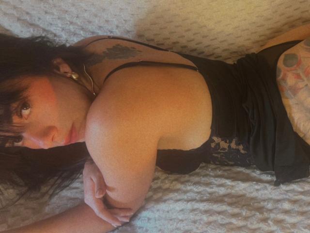 DominatrixMelissa ligt op een bed, ze draagt een zwarte kanten top, haar billen zijn bloot, ze heeft een tatoeage aan de rechterkant en kijkt terug met rode wangen..