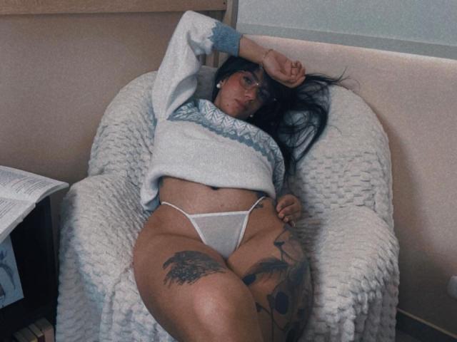 Dominatrix Melissa liegt auf einem Stuhl, trägt einen grauen Pullover und einen weißen String, der ihr Gesäß und ihre Vulva entblößt. Sie hat Tattoos an ihrem Oberschenkel und Arm, sowie langes, schwarzes Haar..