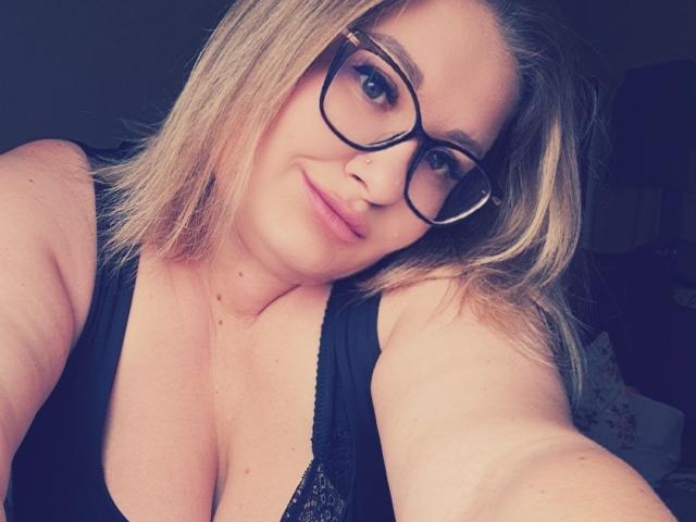 RadaRori with blonde hair, glasses, and a nose ring, showing cleavage in a black lace top, smiling slightly. Her tits are visible, she's taking a selfie.