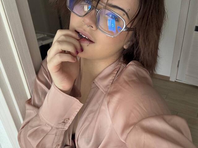 BarrieGromley with glasses, nose piercings, and a lip piercing, wearing a pink satin shirt, biting her finger, showing a cute, sexy face, brown hair, and light skin.