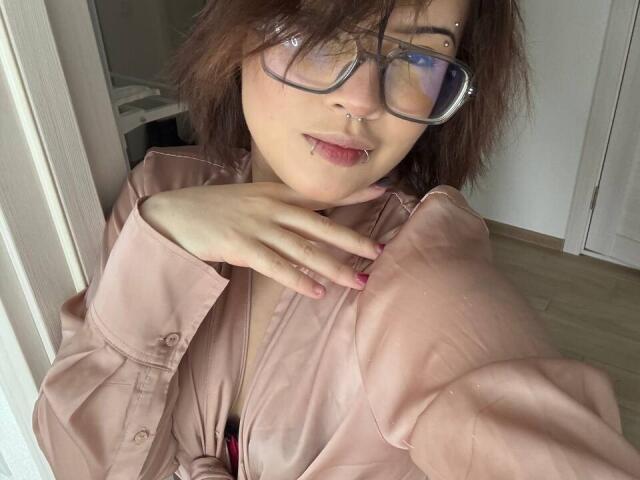 BarrieGromley with glasses, nose piercings, and lip piercing, wearing a pink button-up shirt, hand on chest, pink nails, in a bright room, wood floor, and white walls.