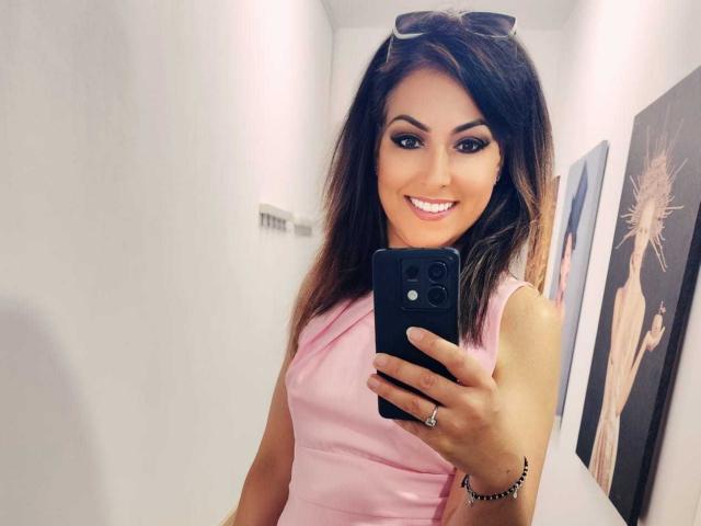 LisaCroftt, com cabelo castanho, vestindo um vestido rosa, segurando um telefone preto, sorrindo para a câmera, paredes brancas e obras de arte ao fundo. Suas nádegas não estão visíveis..