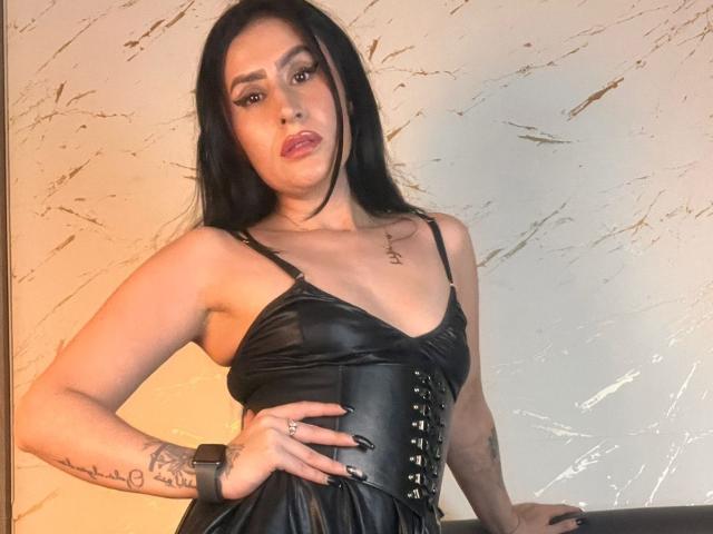DemiMistral, avec ses longs cheveux noirs, porte une robe en cuir noir, un corset à lacets et des ongles noirs. Elle a des tatouages sur son bras droit et porte une montre. Sa poitrine est visible..