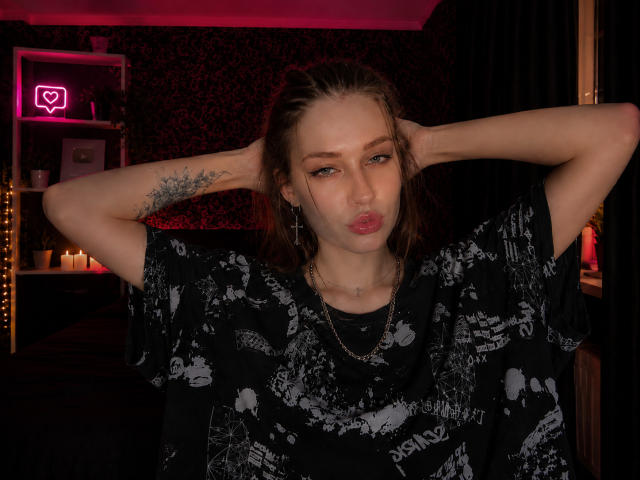 MechelleCordy with pouty lips, black graphic tee, arms raised, tattoo on left arm, brown hair in braids, pink eye makeup, gold necklace, in dimly lit room.