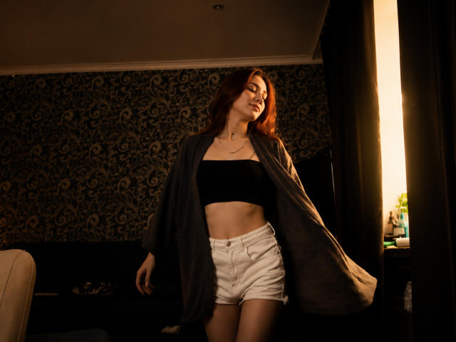 MechelleCordy with red hair, black strapless top, white shorts, and gray open shirt, standing in dimly lit room, smiling, showing her flat abs and small tits.