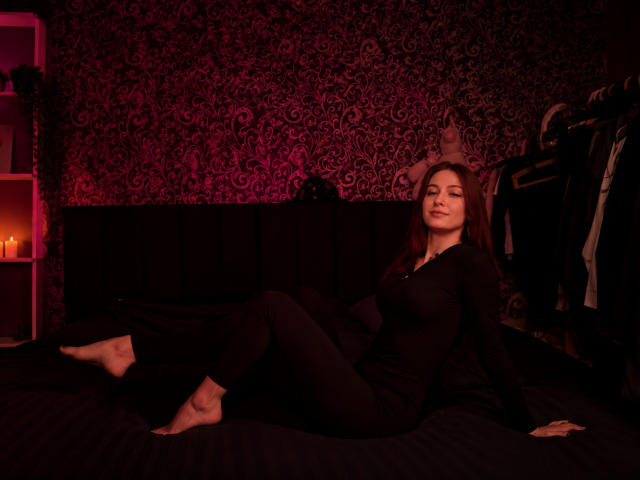 MechelleCordy, barefoot, in black outfit, sitting on dark bed, legs up, arms back, red patterned wall, candles in shelf, clothes on right, soft red lighting, relaxed expression.
