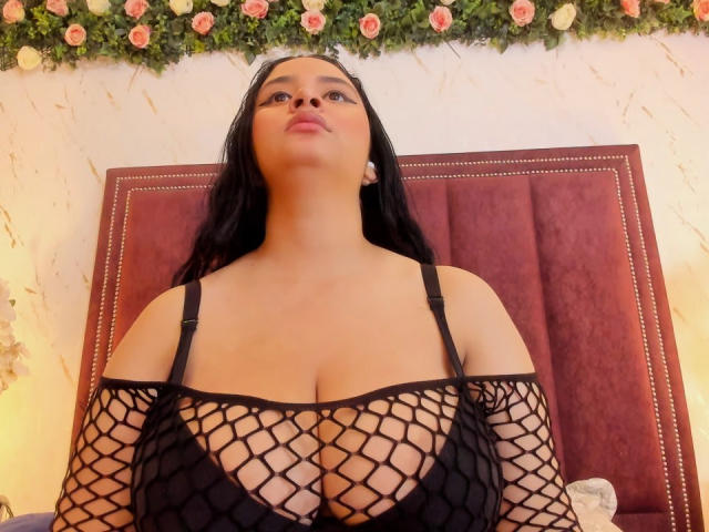 GraceBlue69 con grandi seni, indossa una maglia nera a rete, guarda verso l'alto, ha lunghi capelli neri, orecchini e in secondo piano c'è una ghirlanda di fiori rosa. È seduta su una sedia di velluto rosso..