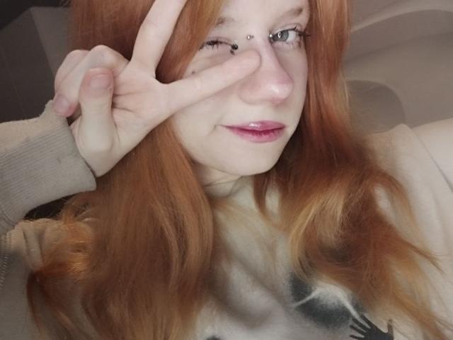 JamieLathrum with red hair, making a peace sign over her right eye, wearing a white shirt, has pale skin, and glossy pink lips, in a selfie.