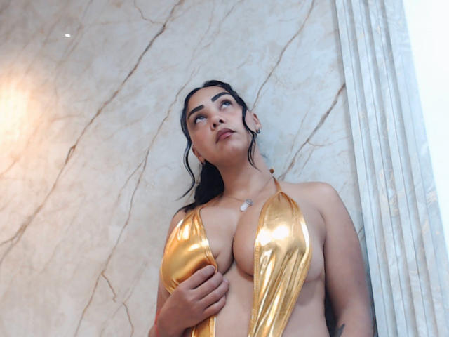 Melany Contrera in einem glänzenden goldenen Bikini, der sie benutzt, um ihre großen, runden Brüste zu bedecken, blickt mit dunklem Haar, heller Haut und einem subtilen, selbstbewussten Ausdruck nach oben..