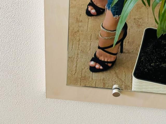 Trima's schwarze Sandaletten mit hohen Absätzen und goldenen Ketten an den Knöcheln, die weiß lackierte Zehen zeigen, stehend auf einem beige gefliesten Boden, reflektiert in einem Spiegel mit einer Zimmerpflanze..