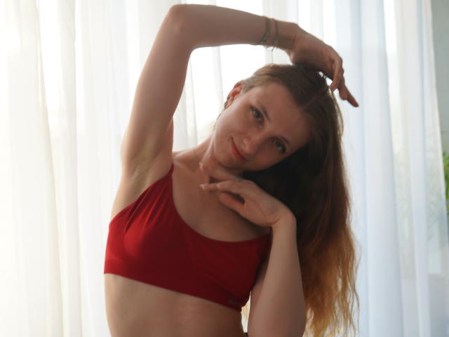 DeandreaLowrance with long red hair, wearing a red bra, arms raised, one hand touching her neck, and the other in her hair, smiling at the camera.