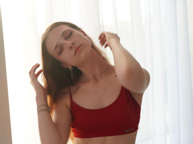 Deandrea Lowrance, con gli occhi chiusi, un reggiseno sportivo rosso e le braccia alzate, ha la pelle chiara, i capelli castani e un piercing al naso, ed è in piedi in una luce soffusa e luminosa..