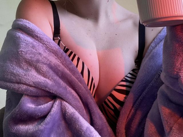 Os seios da FlyFreedom, em um sutiã listrado preto e laranja, cobertos por um robe roxo, felpudo e com um ombro de fora, segurando uma caneca rosa e texturizada. Luz suave e natural..