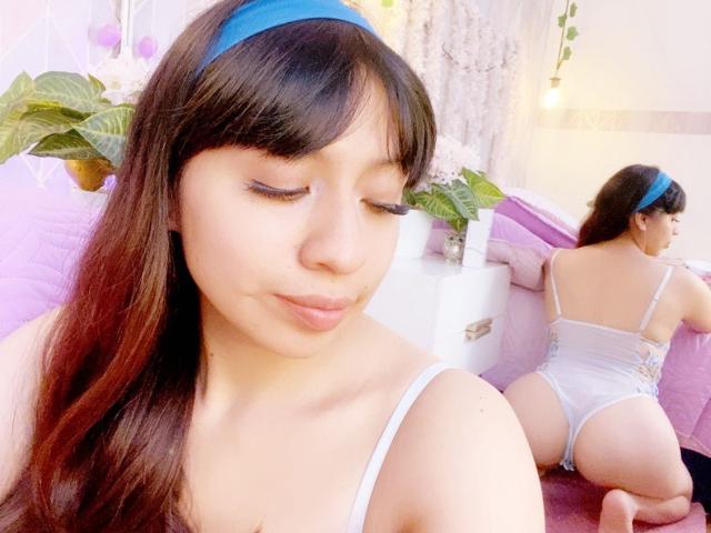 ZoeRedds, con cabello castaño y una diadema azul, con los ojos cerrados, vestida con lencería blanca, mostrando su trasero, arrodillada sobre una cama rosa, reflejada en un espejo. Habitación suave, iluminada en tonos rosados..