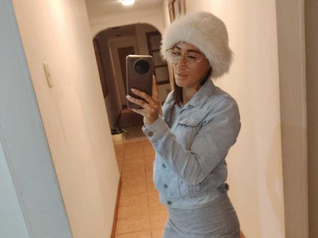 SharissWilliam con un cappello di pelliccia bianca, occhiali e un outfit in denim grigio, che tiene un telefono, mostra un selfie nello specchietto laterale, mostrando un sedere carino, in un corridoio..