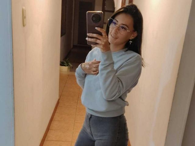 SharissWilliam taking a mirror selfie, wearing glasses, a gray sweater, and high-waisted jeans, with a hand on her chest, smiling slightly, in a hallway.
