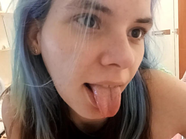 MathaDernier's face, blue-tipped hair, and tongue out. She's sticking her pink tongue out, showing her small nose and light skin. She has a slight, cute, and playful expression.