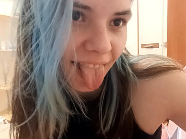 MathaDernier mit blau gefärbten Haaren, die die Zunge herausstreckt und einen niedlichen Gesichtsausdruck hat, trägt ein schwarzes Oberteil, das ein nacktes Schulter zeigt. Sie hat helle Haut und hellbraue Augenbrauen..