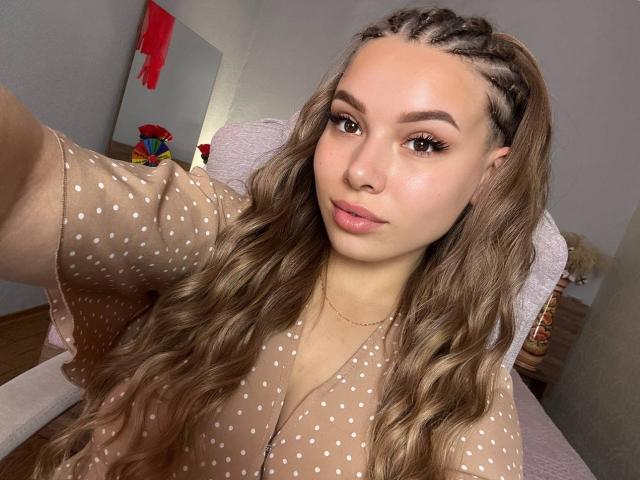 Niferttiti has long, wavy brown hair with braids, wearing a beige polka dot top, looking at the camera with full lips and a slight smile, holding a phone.