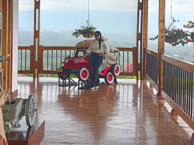 TamaraFoxy sentada com o traseiro em um jipe de brinquedo vermelho, chão de madeira, plantas penduradas e vista para as montanhas, vestindo uma jaqueta bege e calças pretas, com uma bolsa nas costas..