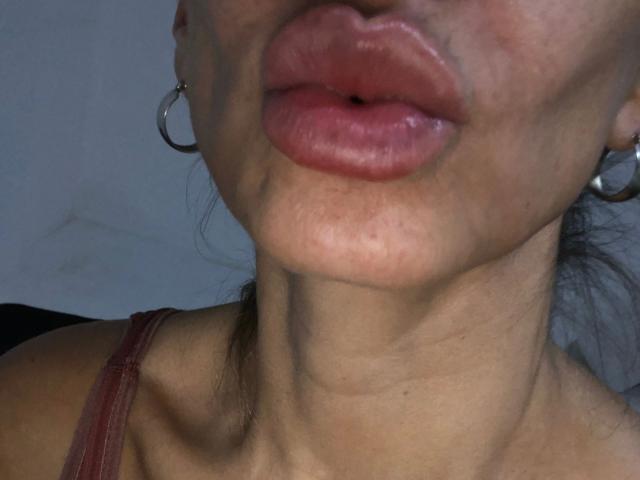 Os lábios volumosos, rosados e marrons de MyrellaPink-hot estão ligeiramente entreabertos, revelando um pequeno orifício escuro no centro. Ela está usando brincos de argola prateados e uma alça vermelha..