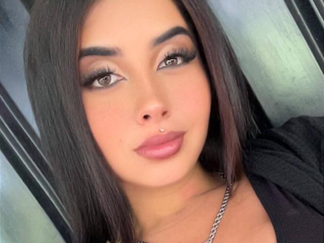 ValerySilva has dark brown straight hair, thick eyebrows, long eyelashes, brown eyes, a nose piercing, and pink glossy lips. She's wearing a black top with a silver necklace.