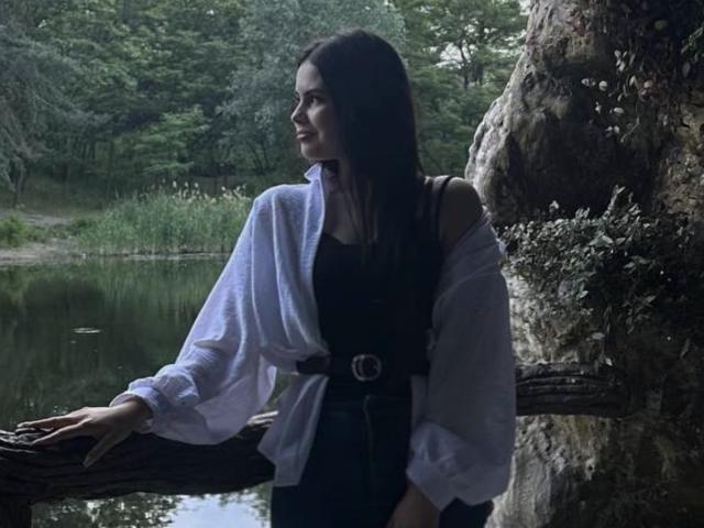 JennaMiu está de pie junto a un árbol, vestida con una camisa blanca con un hombro descubierto, un vestido negro y un cinturón, con el pelo largo y oscuro, mirando hacia un lado, cerca de una fuente de agua reflectante..