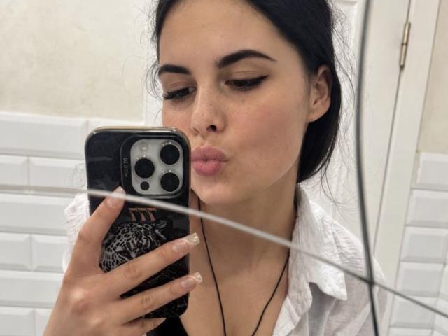 JennaMiu, con le labbra piene, le sopracciglia scure e i capelli neri, tiene in mano un telefono con una custodia a stampa leopardo, indossa una camicia bianca e si guarda allo specchio..