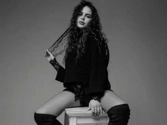 JennaMiu com cabelo cacheado, blusa preta, mangas de renda, sentada em um banquinho, pernas abertas, segurando o cabelo, usando botas que vão até a coxa, shorts pretos, com um sorriso malicioso, foto em preto e branco..