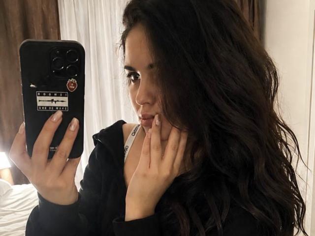 JennaMiu, com cabelos castanhos longos e ondulados, segurando um telefone preto, vestindo uma jaqueta preta e cobrindo a boca com a mão, mostrando uma selfie no espelho..