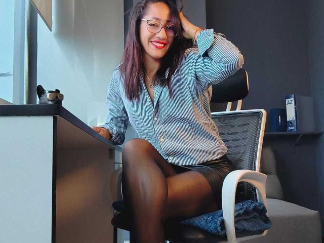 SamanthaCorine, mit rotem Lippenstift und Brille, sitzt auf einem Stuhl, trägt ein gestreiftes Hemd, einen schwarzen Rock und schwarze Strumpfhosen, und ihre Gesäßpartie befindet sich auf dem Stuhl. Sie lächelt in die Kamera..