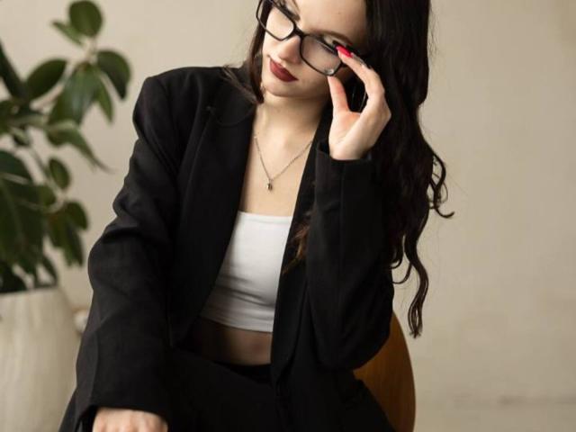Robin Sliwinski, con cabello oscuro y ondulado, gafas negras, uñas rojas, un blazer negro, una camiseta corta blanca, una falda marrón, tocándose las gafas, llevando un collar y de pie junto a una planta verde..