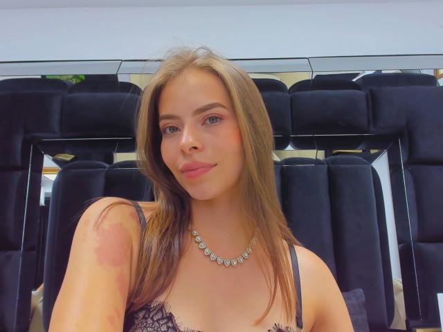 AnnaFer with brown hair, blue eyes, and pink lipstick, wearing black lace lingerie, necklace, and a bruise on her right shoulder, smiling in a car with black seats.