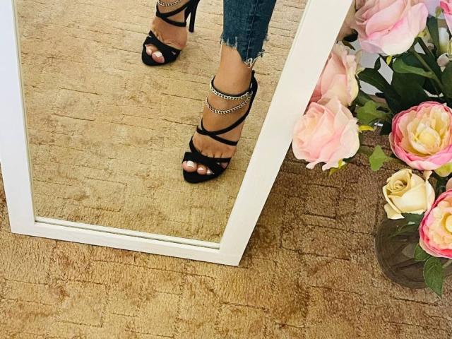 Los tacones altos negros con tiras de Trima, con detalles de cadena plateada, mostrando sus dedos de los pies pintados, se reflejan en un espejo blanco, mientras está de pie sobre un suelo de madera con rosas rosadas cerca..